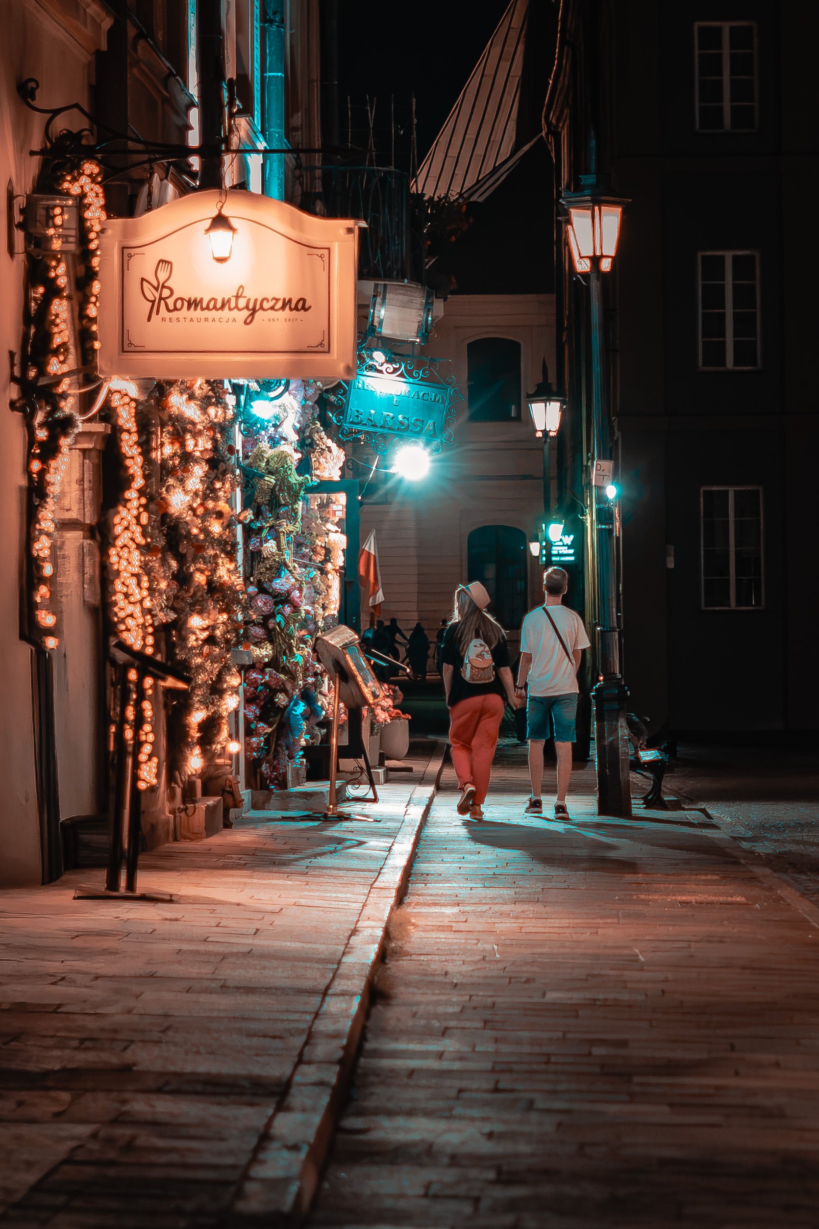 A couple walking a cobbled old town street past a lit restaurant at night