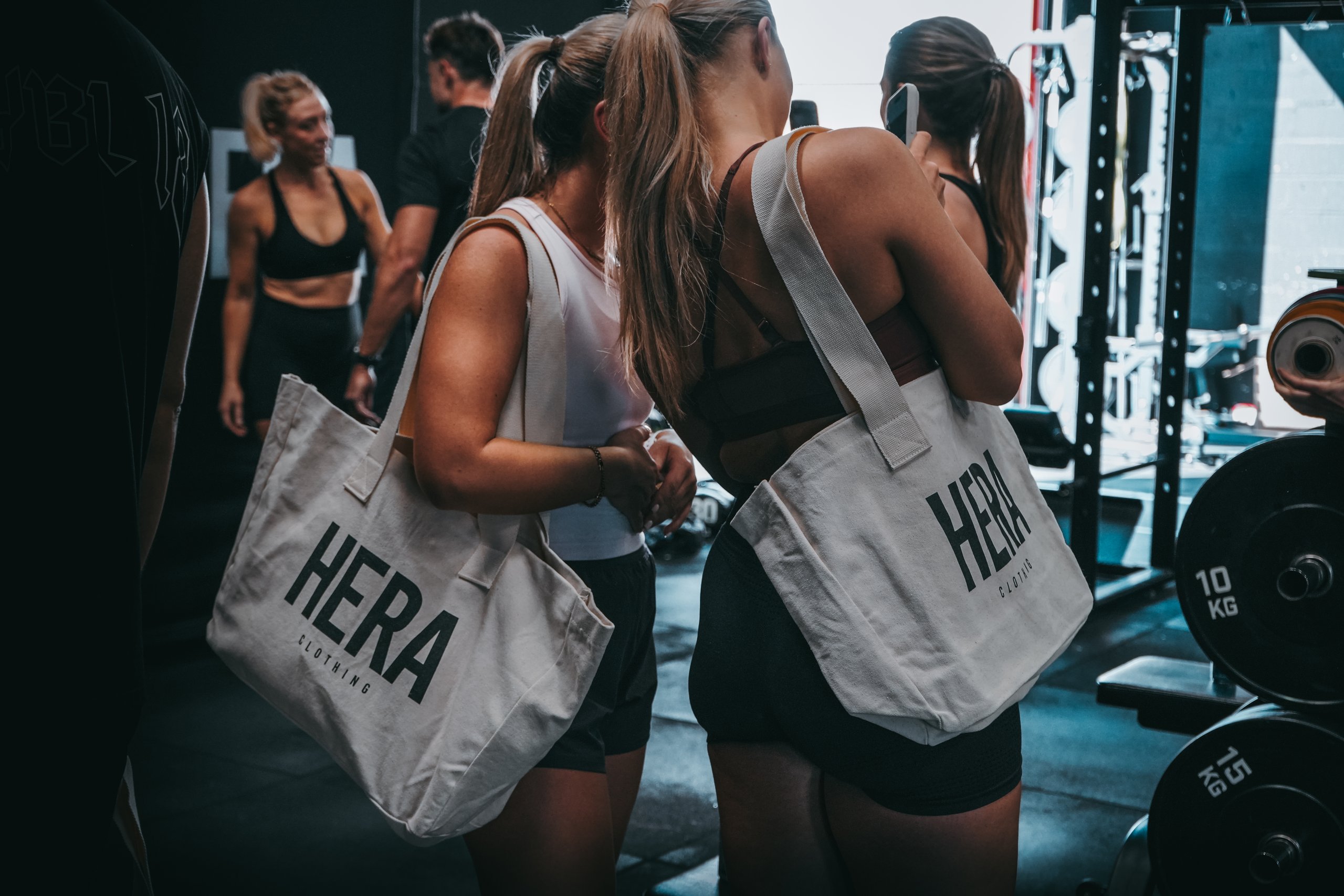 Two women carrying HERA Clothing tote bags at a gym event