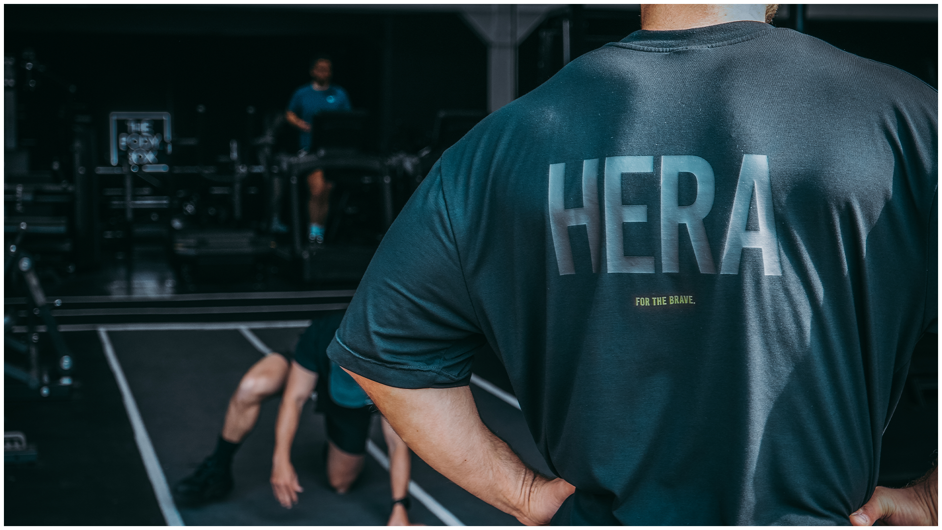 A man in a HERA training top watching an athlete work out on a gym floor