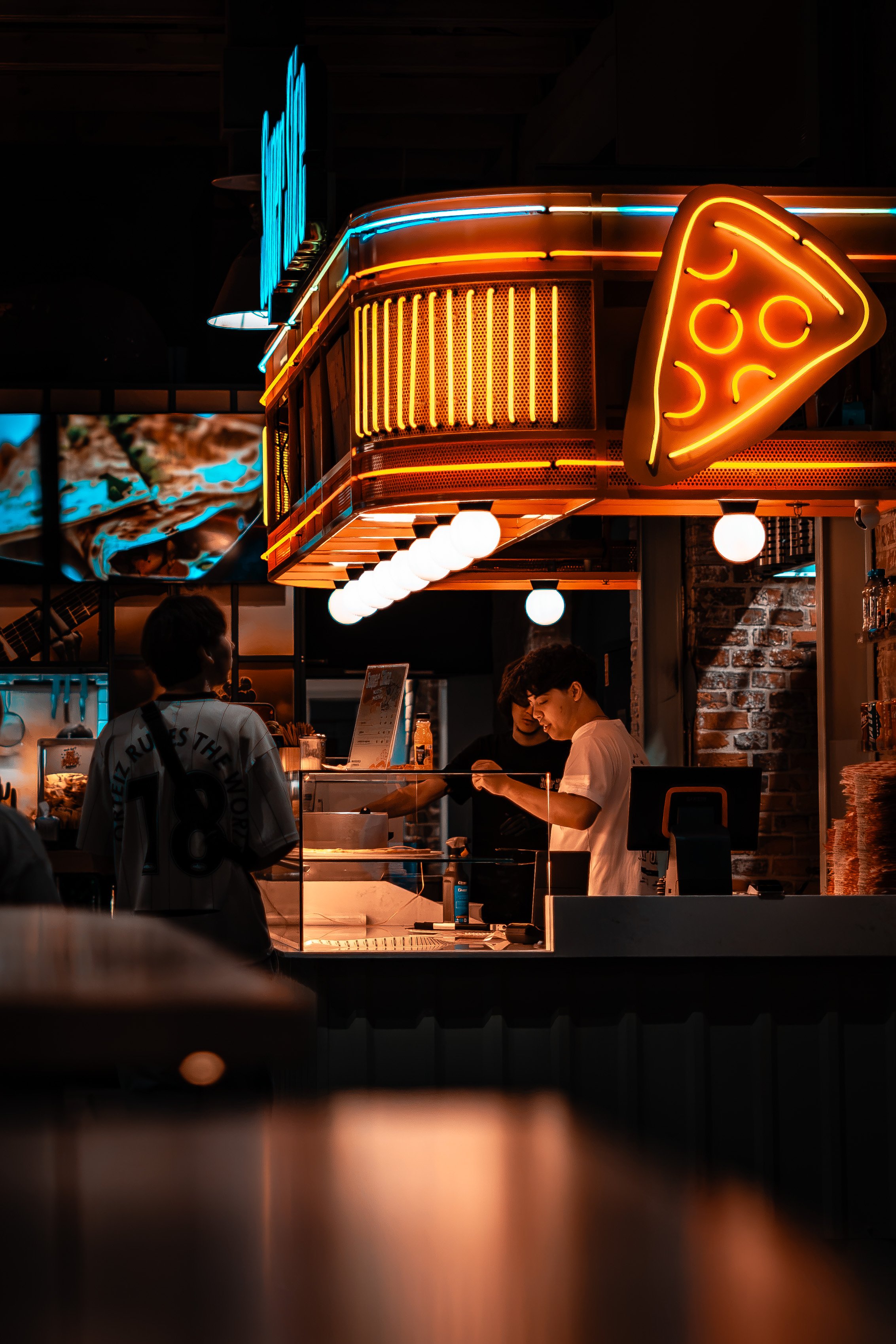 Staff serving behind a pizza counter under a neon slice sign at night