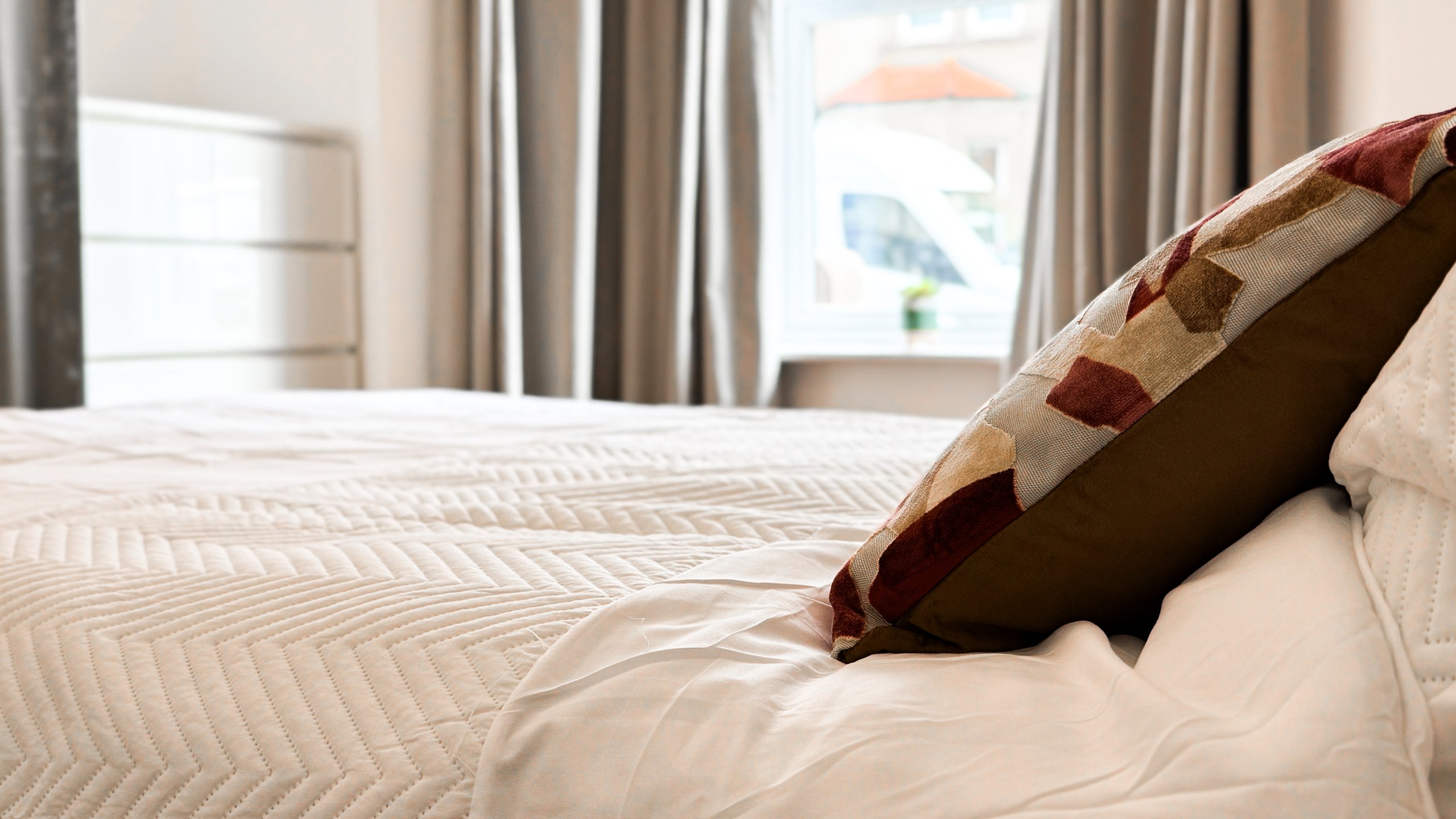 A made bed with a patterned cushion beside a bright bedroom window